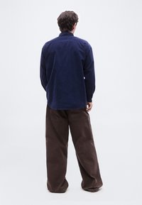 Un homme se tient de dos, portant une chemise à manches longues bleu foncé et un pantalon brun ample, sur un fond blanc uni.