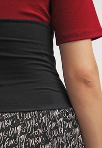 Punainen paita lyhyillä hihoilla, musta vyötäröosa ja kuvioitu hame, jossa on valkoista käsinkirjoitettua tekstiä mustalla taustalla.