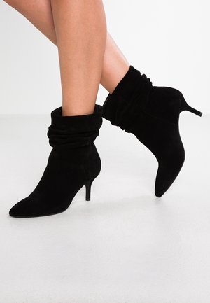 Jambes portant des bottines en daim noir décontractées avec des bouts pointus et des talons kitten, sur un fond blanc uni.