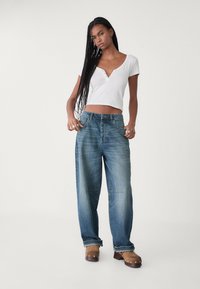 Lättviktig vit kortärmad topp med v-ringning, tillsammans med löst sittande blå denimjeans och bruna träskor med metalliska detaljer.