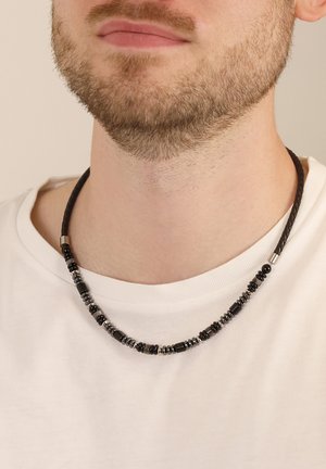 Schwarze Perlenkette mit glatten und strukturierten Elementen, die zylindrische und runde Formen aufweist und über einem weißen Hemd getragen wird.