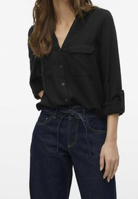 Camicia nera con colletto a V, due tasche sul petto, maniche arrotolate, abbinata a jeans blu scuro con cintura a cravatta.