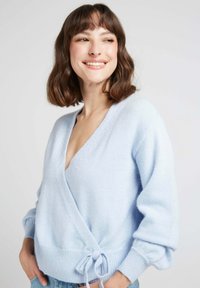 Pull en maille tricotée douce de couleur bleu clair, avec un décolleté en V profond, des manches larges et un accent à la taille avec un nœud.