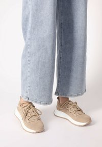 Beżowe sneakersy z teksturowanym zamszowym wierzchem, zawiązanym sznurowaniem i białą podeszwą środkową. Noszone z jasnoniebieskimi spodniami w szerokim kroju.