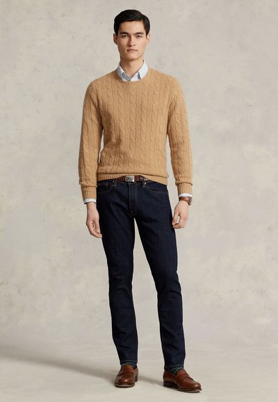 Joven de pie, vestido con un suéter de punto trenzado color tostado, camisa azul claro con cuello, vaqueros oscuros, cinturón marrón, reloj y zapatos de cuero marrón.