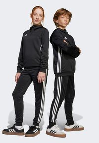 Zwarte sportieve trainingspakken met witte strepen aan de zijkanten. Het meisje draagt een capuchon, beiden dragen contrasterende schoenen met rubberen zolen.