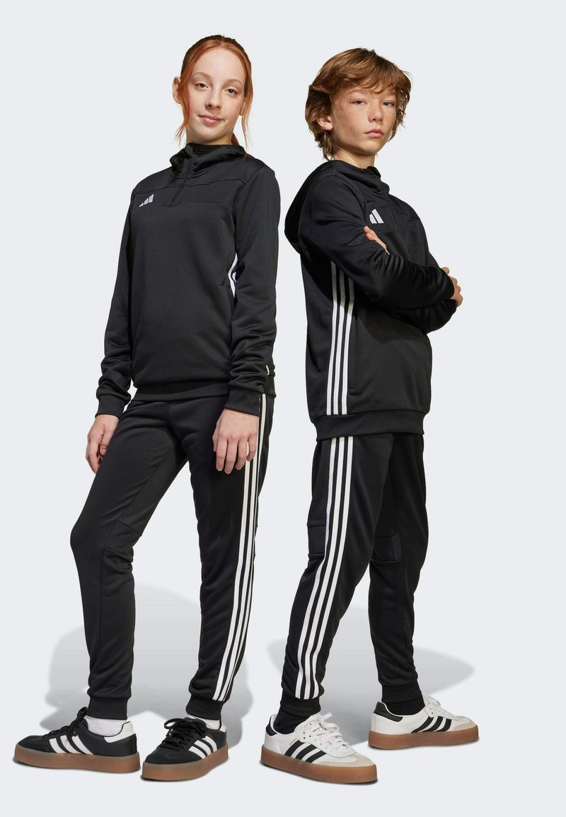Zwarte sportieve trainingspakken met witte strepen aan de zijkanten. Het meisje draagt een capuchon, beiden dragen contrasterende schoenen met rubberen zolen.