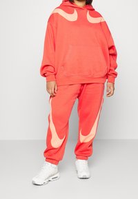 Korallfärgad hoodie med stor rosa swoosh-design och framficka; matchande sweatpants med swoosh-accenter på sidorna. Vita sneakers.