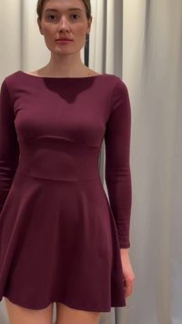 Γυναίκα που φοράει εφαρμοστό, μακρυμάνικο, μπορντό φόρεμα, στέκεται μπροστά από κουρτίνες ανοιχτού χρώματος, κοιτάζοντας μπροστά με ουδέτερη έκφραση.