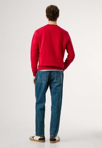 Hombre de pie de espaldas, vistiendo un sudadera roja, jeans azules y zapatillas blancas con detalles rojos y negros, sobre un fondo simple.