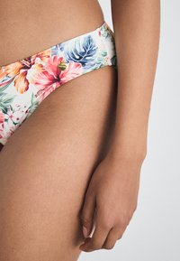 Blomstrete bikinitruse med lys bakgrunn, som har livlige rosa og blå tropiske mønstre og en liten gulllogo på linningen.