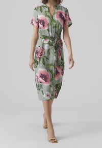 Donna che cammina in un abito midi bottonato con grande stampa floreale rosa, maniche corte e una cintura annodata in vita, indossando sandali open-toe nudi.