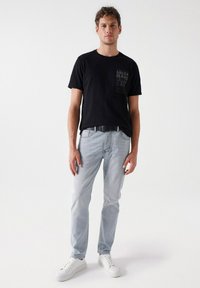 Camiseta negra con un estampado texturizado del logo en el pecho, combinada con jeans de color azul claro y zapatillas blancas. Ajuste casual y diseño sencillo.