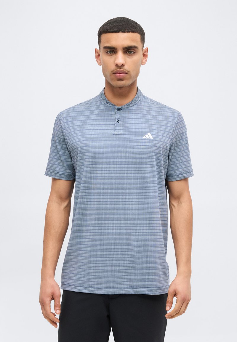 Νεαρός άνδρας που φοράει κοντομάνικο, ανοιχτό μπλε πόλο με σχέδιο και μικρό λευκό λογότυπο στο αριστερό στήθος, στέκεται μπροστά σε ένα μονόχρωμο φόντο.