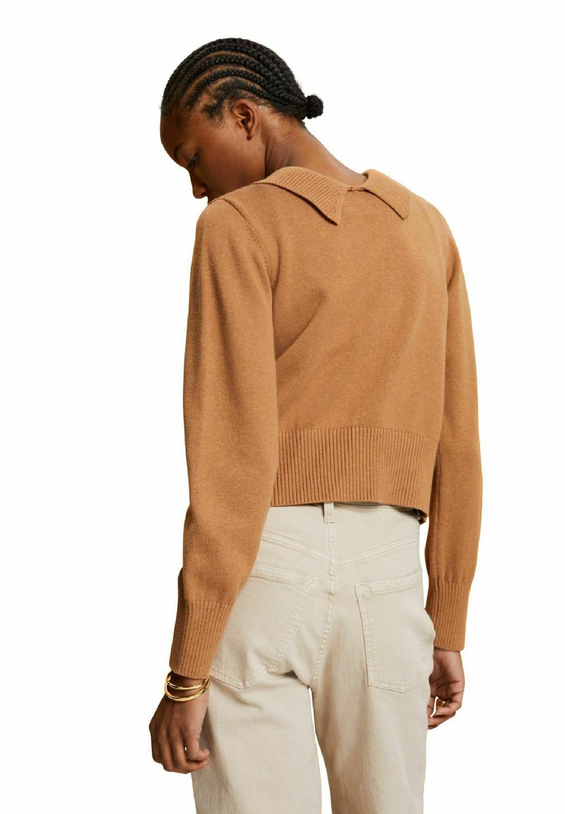 Pull en cache-cœur marron avec un ourlet et un col côtelés. Le modèle porte un pantalon taille haute de couleur claire. Texture tricotée avec des manches longues.