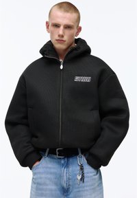 Fiatal férfi rövid szőke hajjal, fekete cipzáras kapucnis pulóvert visel, amelyen "STUDIO" logó látható, kezét a zsebében tartja, világoskék farmert visel, amelyhez kulcsok vannak csatolva.