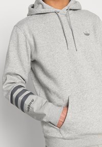 Šedá bavlněná mikina s klokaní kapsou, kapucí se šňůrkou a třemi šedými pruhy na levém rukávu. Na hrudi je vyšitý logo.