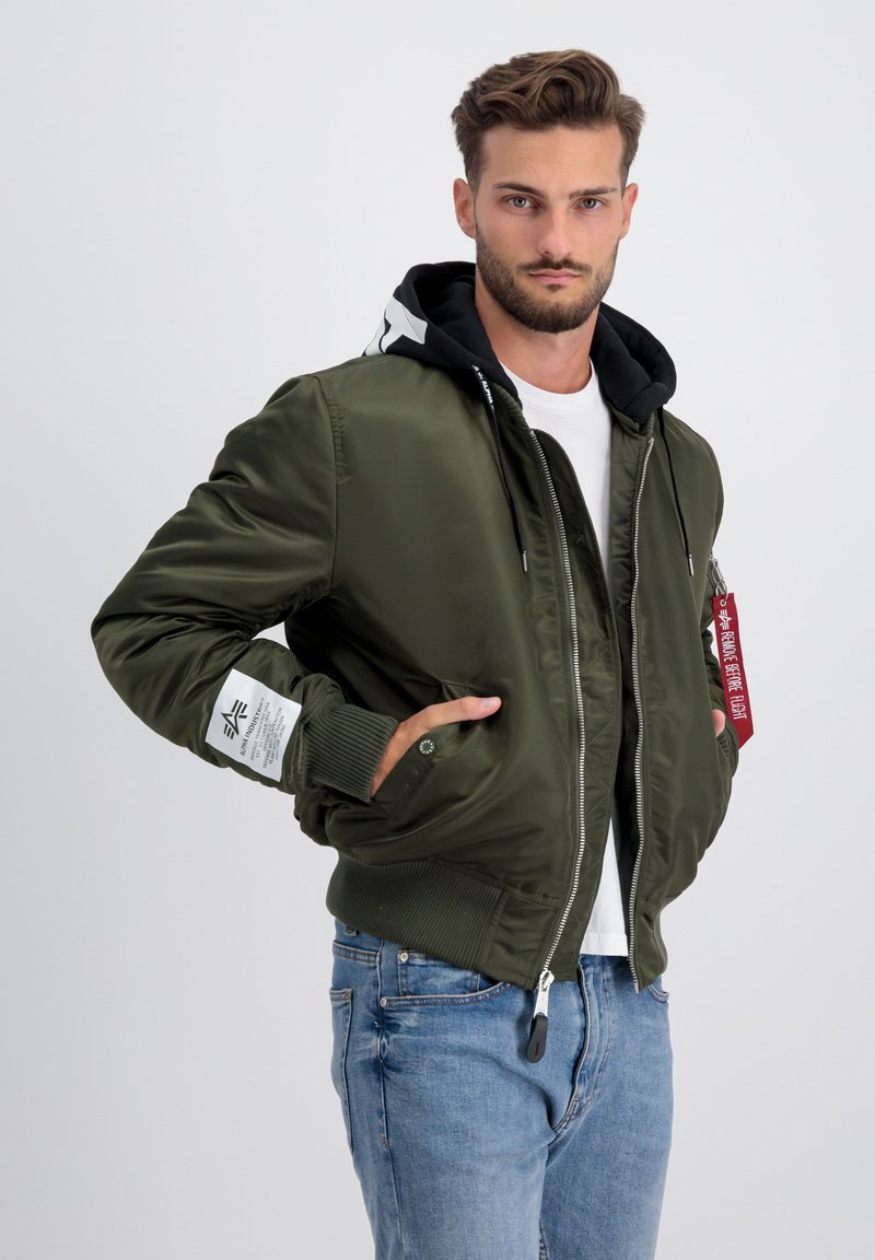 Olivnozelen bomber jakna s črno kapuco, belimi detajli, rebrastimi manšetami in zadrgo spredaj. Ima rdečo etiketo in žep na rokavu.