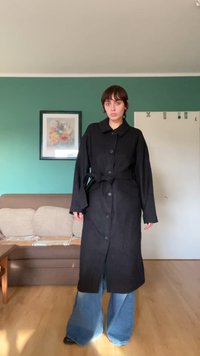 Black oversized coat with a collar, front buttons, and a tie waist. Loose-fitting sleeves and two side pockets. Textured fabric.
