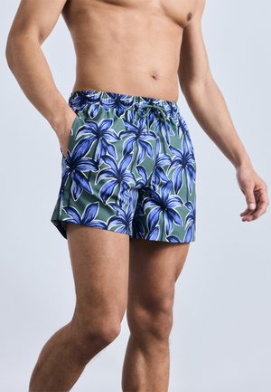 Mand stående med den ene hånd i lommen iført grønne badeshorts med blåt blomsterprint og snøre i taljen mod ensfarvet baggrund.