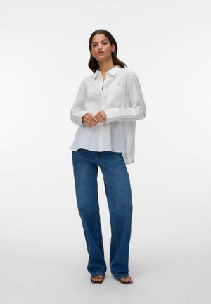 Kvinde stående iført en løs hvid langærmet knap-på-skjorte, blå vidde-ben-jeans og åbne tåsandaler mod en ensfarvet baggrund.