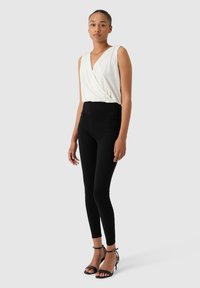 Femme debout portant un haut enveloppant sans manches blanc, des leggings noirs taille haute et des escarpins à bride de cheville noirs sur un fond gris uni.