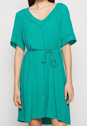 Woman wearing short teal dress with V-neck, short sleeves, decorative stitching, and a tied waist belt, standing against a plain background.