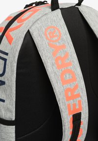 Mochila de tela gris con correas acolchadas, que presenta un llamativo logo en naranja "SUPERDRY®" y un panel trasero de malla negra.