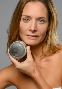 Latta rotonda in metallo argentato di "Poudre de Riz Dermophile" con testo in rilievo, tenuta vicino al viso di una persona; superficie liscia, dettaglio decorativo sul bordo.