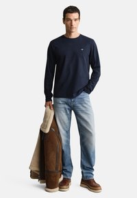 Marineblå langærmet skjorte, lyseblå jeans, brune læderstøvler og en brun jakke foret med lammeskind. Enkel logo på skjorten.