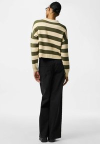 Osoba z włosami upiętymi w koka, nosząca okulary, oliwkowo-beżowy sweter w paski oraz czarne szerokie spodnie, stojąca na tle jednolitej białej ściany.