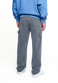 Man som bär lösa grå cargobyxor med stora bakfickor, en blå sweatshirt och vita sneakers, sedd bakifrån på en vit bakgrund.