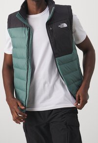 Grüner und schwarzer gepolsterter Weste mit hohem Kragen, Reißverschluss und Logodetail auf der Brust. Hergestellt aus leichtem, strukturiertem Stoff.