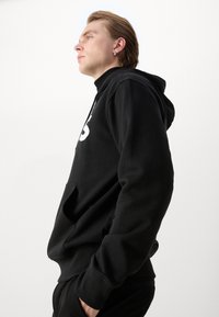 Jonge persoon met licht haar, draagt een zwarte hoodie met witte opdruk, handen in de zakken, kijkt zelfverzekerd naar links op een effen witte achtergrond.