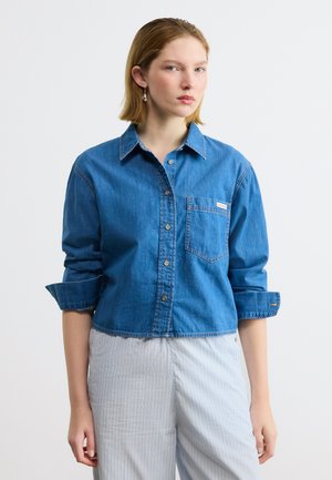 Jeune femme portant une chemise en jean bleue à boutons avec les manches retroussées et un pantalon clair à fines rayures, debout devant un fond uni.