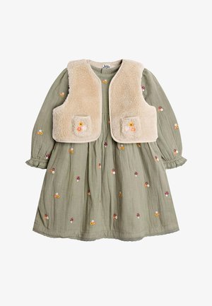Lächelndes junges Mädchen mit langen dunklen Haaren, das ein grünes Kleid mit Blumenstickerei und eine beige Fleeceweste trägt, draußen in einem bewaldeten Gebiet.