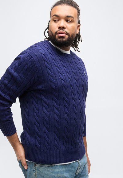 Άνδρας με γένια και πλεγμένα μαλλιά, ντυμένος με μπλε πουλόβερ με πλεκτό σχέδιο και τζιν, κοιτάζει προς τα αριστερά του, μπροστά σε λευκό φόντο.