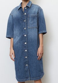 Sukienka koszulowa z denimu w średnim niebieskim kolorze, z krótkim rękawem, z zapięciem na guziki z przodu oraz kieszonką na piersi. Charakteryzuje się wyrazistym przeszyciem i luźnym krojem.