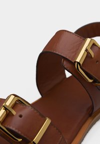 Καφέ δερμάτινο σανδάλι με δύο λουριά και χρυσές ορθογώνιες μεταλλικές αγκράφες, εμφανιζόμενο σε κοντινή λήψη πάνω σε λευκό φόντο.