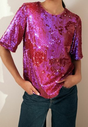 Shiny purple and red sequin short-sleeve top worn with dark blue jeans, hands in pockets, torso visible, neutral background.