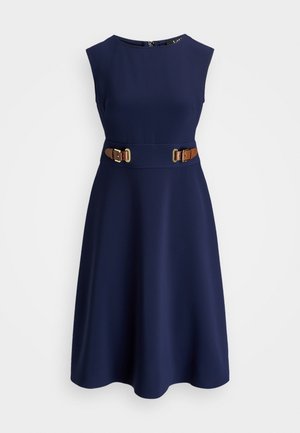 Mouwloze marineblauwe jurk met een aangesloten taille met twee bruine leren gespsluitingen, uitlopende rok en een ronde halslijn.