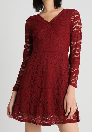 Femme portant une robe en dentelle bordeaux à manches longues avec un col en V et une taille cintrée, affichant un motif floral en dentelle sur toute la robe.