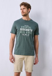 Mörkgrön bomulls-T-shirt med fet vit tryckt text. Korta ärmar, rund halsringning, bärs med ljusbeiga uppvikta shorts. Enkel, avslappnad design.