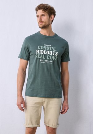 Dark green cotton t-shirt with bold white printed text. Short sleeves, round neckline, worn with light beige rolled shorts. Simple, casual design.