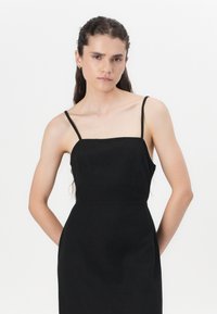 Robe noire sans manches avec de fines bretelles, un décolleté carré et une silhouette ajustée. Texture lisse, design minimaliste, sans motifs ni accents.