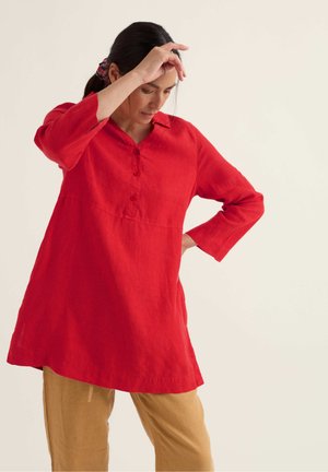 Frau mit zurückgebundenem dunklem Haar trägt eine lange rote Tunika und beigen Hosen, hebt eine Hand zur Stirn und legt die andere auf die Hüfte.