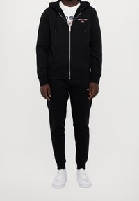 Černá mikina na zip s kapsami a stahovacími šňůrkami, doplněná o odpovídající černé tepláky, nošená s bílými teniskami s hladkým designem.