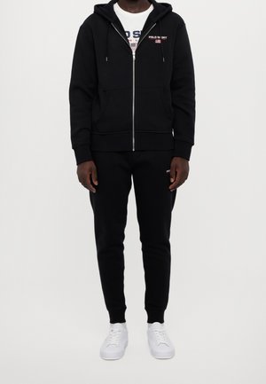 Felpa nera con cerniera, tasche e stringhe, abbinata a pantaloni jogger neri coordinati, indossata con sneaker bianche dal design semplice.