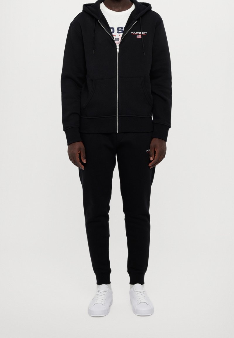 Černá mikina na zip s kapsami a stahovacími šňůrkami, doplněná o odpovídající černé tepláky, nošená s bílými teniskami s hladkým designem.