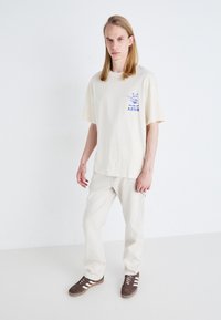 Tricou oversized crem cu grafică imprimată albastră, asortat cu pantaloni bej deschis și adidași maronii cu dungi albe. Textură de țesătură netedă.